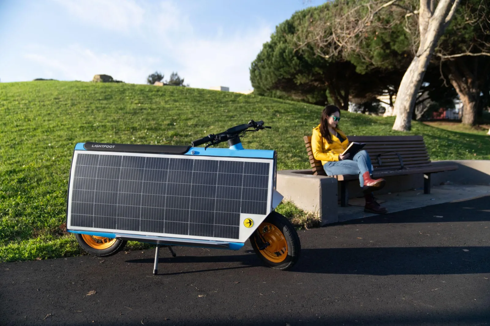 A Smile Machine parked on a path with its solar panel facing the sun while its rider reads on a park bench
