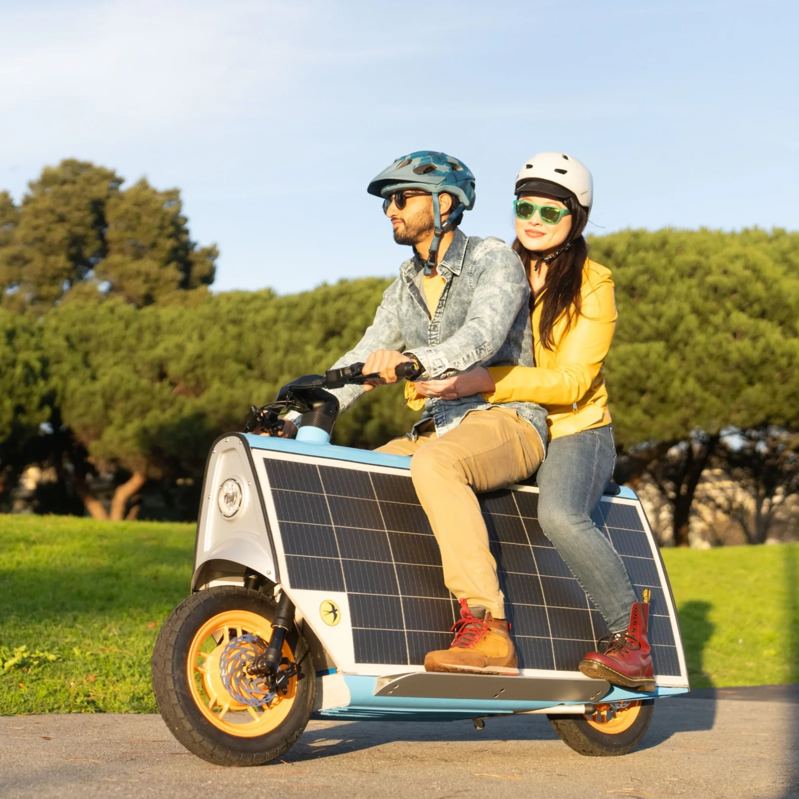 Two riders in helmets and sunglasses on a sky blue Smile Machine, rolling through a sunlit park