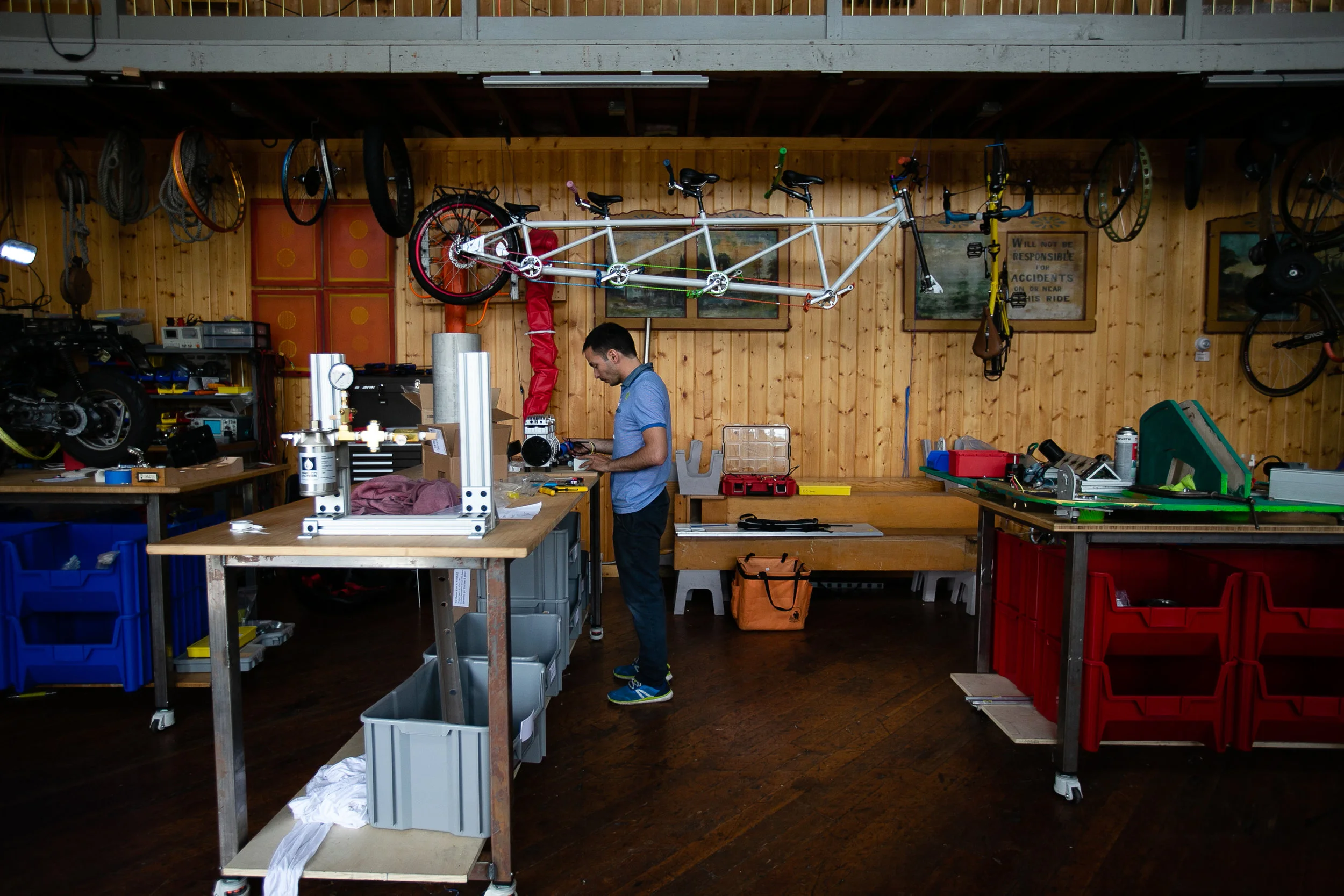 The Otherlab workshop: an engineer at a workbench beneath a tandem bicycle frame and bike wheels hung from a wood-panelled wall, with prototype parts and tool bins around the room.